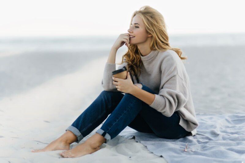 Photo d'une jeune femme assise sur le sable regardant l'horizon perdue dans ses pensées.