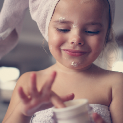 Photo d'une petite fille plongeant son index dans un pot de crème et l'étalant sur son visage délicat.